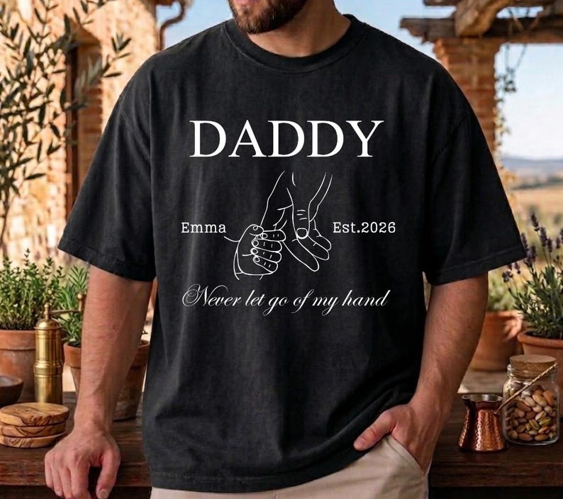 May include: Black t-shirt with the word "DADDY" in white block letters. Below the word "DADDY" is a graphic of two hands holding each other, with the name "Emma" and the date "Est.2026" printed on the shirt. The phrase "Never let go of my hand" is printed below the graphic.