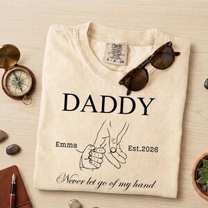 May include: Cream-colored t-shirt with the word "DADDY" in black, a graphic of two hands holding, and the text "Emma Est. 2026 Never let go of my hand." A pair of sunglasses rests on the shirt.
