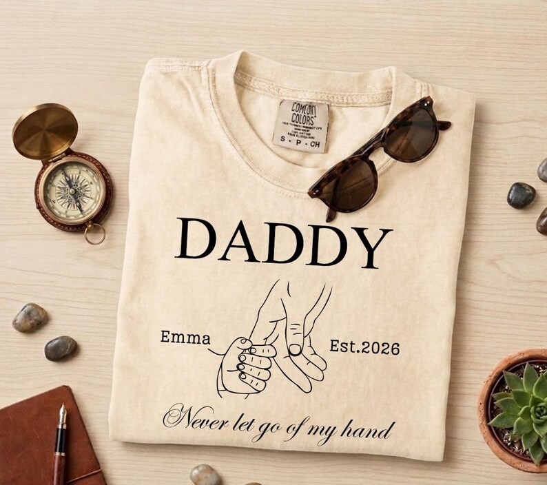 May include: Cream-colored t-shirt with the word "DADDY" in black, a graphic of two hands holding, and the text "Emma Est. 2026 Never let go of my hand." A pair of sunglasses rests on the shirt.
