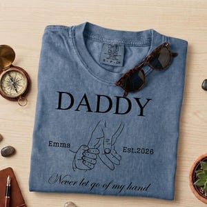 May include: A blue Comfort Colors t-shirt with the word "DADDY" printed in black. Below the word is a graphic of two hands holding each other, with the text "Emma" and "Est. 2026". The phrase "Never let go of my hand" is printed at the bottom. Sunglasses rest on the shirt.