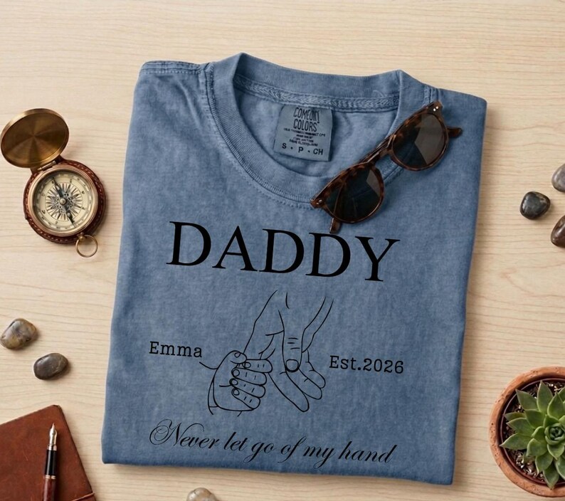 May include: A blue Comfort Colors t-shirt with the word "DADDY" printed in black. Below the word is a graphic of two hands holding each other, with the text "Emma" and "Est. 2026". The phrase "Never let go of my hand" is printed at the bottom. Sunglasses rest on the shirt.
