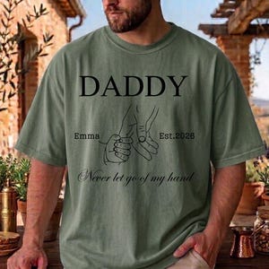 May include: Olive green t-shirt with the word "DADDY" in large black letters. Below, a line drawing depicts a hand holding a smaller hand, with the text "Emma Est. 2026" and "Never let go of my hand".