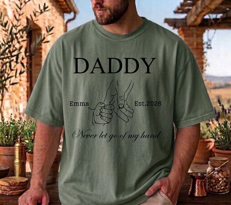 May include: Olive green t-shirt with the word "DADDY" in large black letters. Below, a line drawing depicts a hand holding a smaller hand, with the text "Emma Est. 2026" and "Never let go of my hand".