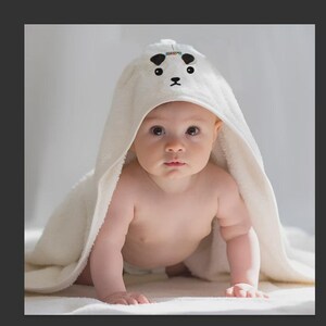 Puede incluir: Un bebé con una toalla con capucha blanca con un diseño de cara de panda. El bebé está gateando sobre una superficie blanca, mirando directamente a la cámara. La toalla tiene ojos negros y una nariz negra.