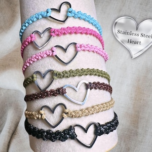 May include: A collection of macrame bracelets in various colors, each featuring a silver heart charm. The bracelets are displayed on a white cylindrical form. The colors include blue, pink, green, brown, tan, and black. The text "Stainless Steel Heart" is visible.