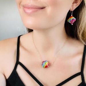 Könnte beinhalten: Eine goldene Halskette mit einem Regenbogen-farbenen, zehnseitigen Würfelanhänger. Passende Regenbogen-Würfelohrringe sind ebenfalls zu sehen. Der Schmuck wird mit einem schwarzen Tanktop getragen. Der Würfel hat weiße Zahlen.