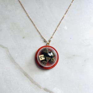 Op de afbeelding: Een goudkleurige ketting met een hanger. De hanger is een heldere, cirkelvormige behuizing met een rood houten frame, met daarin verschillende dobbelstenen. De dobbelstenen zijn zwart met goudkleurige markeringen. De ketting heeft kleine witte kralen bij de sluiting.