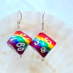 Könnte beinhalten: Ein Paar Ohrringe in Form von Regenbogenwürfeln. Jeder Ohrring ist ein vielseitiger Würfel mit weißen Zahlen und einem Regenbogenverlauf. Die Ohrringe haben goldfarbene Haken und kleine weiße Perlen.