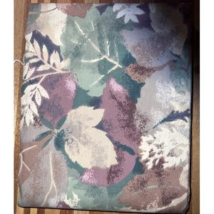 May include: A rectangular cushion with a botanical leaf pattern. The leaves are in shades of green, purple, beige, and brown. The cushion is placed on a wooden surface.