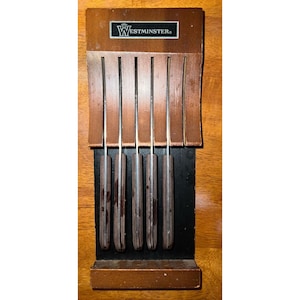 May include: A wooden knife block with six knives. The block is brown with the word "Westminster" printed on the top. The knives have wooden handles and silver blades. The knives are stored in individual slots.