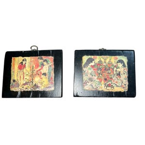 May include: Two vintage framed Asian art pieces. Each features a colorful illustration of people in traditional clothing. The frames are black with a metal loop for hanging. The art depicts scenes of daily life.