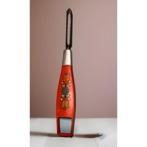 May include: A vintage red vegetable peeler with a floral design. The peeler has a silver blade and handle, and a decorative pattern of flowers in yellow, green, and orange. The peeler is likely made of metal and wood.