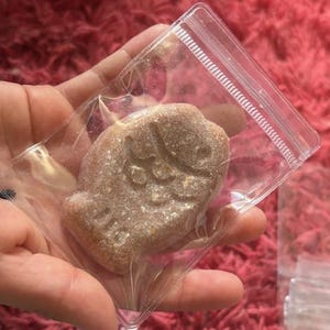 May include: A brown, fish-shaped item is sealed inside a clear plastic bag. The bag is held in a person's hand against a fuzzy, red background. The fish has a textured surface and a detailed design.