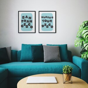 Puede incluir: Dos impresiones art&iacute;sticas enmarcadas con marcos negros cuelgan sobre un sof&aacute; turquesa. Las impresiones tienen un fondo azul claro con las palabras "SAN FRANCISCO" y "WHERE'S YOUR DISCO" en negro. Una mesa de centro redonda de madera y una planta en maceta est&aacute;n frente al sof&aacute;.