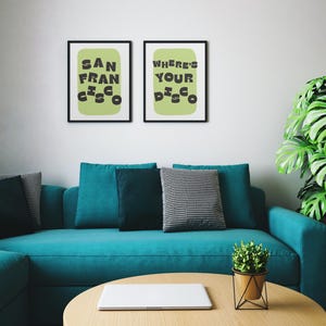 May include: Two framed art prints with black frames hang above a teal sofa. The prints feature the text "SAN FRANCISCO" and "WHERE'S YOUR DISCO" in black on a light green background. A round wooden coffee table and a potted plant are in front of the sofa.