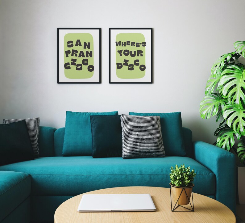 May include: Two framed art prints with black frames hang above a teal sofa. The prints feature the text "SAN FRANCISCO" and "WHERE'S YOUR DISCO" in black on a light green background. A round wooden coffee table and a potted plant are in front of the sofa.