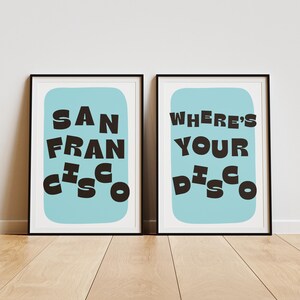 Puede incluir: Dos impresiones art&iacute;sticas enmarcadas con marcos negros. Una impresi&oacute;n dice "SAN FRANCISCO" en letras oscuras y marrones sobre un fondo azul claro. La otra impresi&oacute;n dice "WHERE'S YOUR DISCO" en un estilo similar.