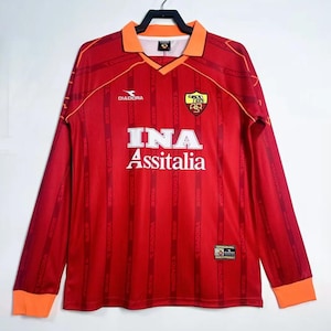 Puede incluir: Una camiseta de fútbol roja de manga larga con detalles naranjas en el cuello y los puños. La camiseta presenta el logotipo "INA Assitalia" y la marca "Diadora". Un escudo del equipo es visible.