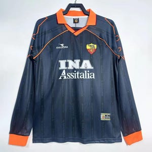 Puede incluir: Una camiseta de fútbol gris oscuro de manga larga con detalles en naranja. La camiseta presenta el logotipo "INA Assitalia" en blanco, un escudo del equipo y el logotipo "Diadora". El cuello y los puños son naranjas.