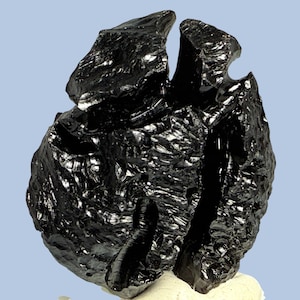 May include: A dark, glossy obsidian stone with a textured surface. The stone has a distinctive shape, featuring a significant split through the middle. The background is a light blue. Obsidian is a natural volcanic glass.
