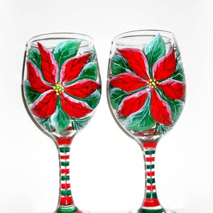 May include: Two clear glass wine glasses with red and green striped stems. Each glass is hand-painted with a red poinsettia flower with green leaves.