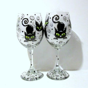 May include: Two clear glass wine glasses with black and white painted designs. Each glass features a black cat with green eyes and a curled tail. The background of the glasses is white with black swirls and dots.
