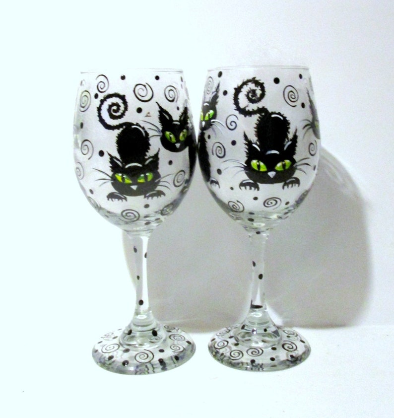 May include: Two clear glass wine glasses with black and white painted designs. Each glass features a black cat with green eyes and a curled tail. The background of the glasses is white with black swirls and dots.