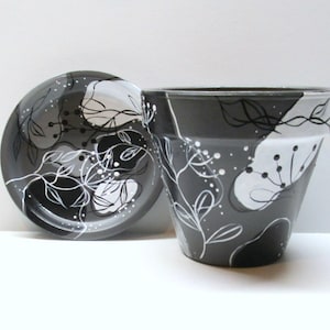 May include: A hand-painted ceramic flower pot and saucer set. The pot and saucer are painted in shades of gray, black, and white, with a floral design. The design features abstract shapes and delicate white lines. The pot is approximately 5 inches tall.