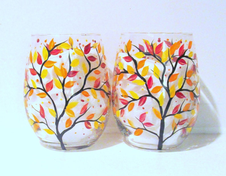 Op de afbeelding: Twee heldere glazen wijnglazen met handgeschilderde herfstbladeren ontwerpen. De glazen hebben een zwarte tak met gele, oranje en rode bladeren.
