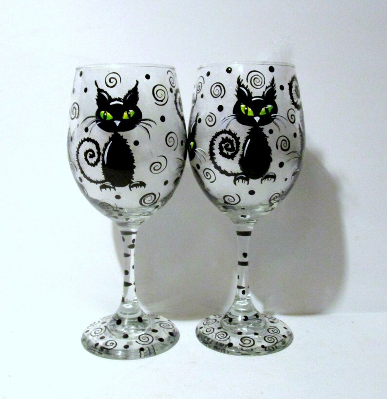 May include: Two clear glass wine glasses with black cat designs. The cats have green eyes and are painted on a white background with black swirls and dots. The glasses have black and white polka dot stems.