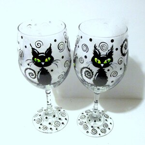 May include: Two clear glass wine glasses with black cat designs. The cats have green eyes and are surrounded by black swirls and dots.