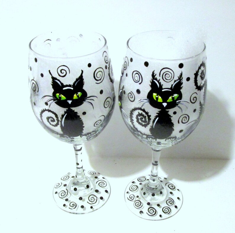 May include: Two clear glass wine glasses with black cat designs. The cats have green eyes and are surrounded by black swirls and dots.