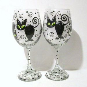 May include: Two clear glass wine glasses with black and white painted designs. Each glass features a black cat with green eyes and a swirling tail. The glasses are decorated with black dots and swirls.