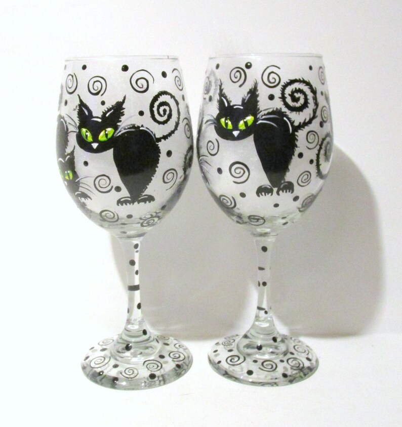 May include: Two clear glass wine glasses with black and white painted designs. Each glass features a black cat with green eyes and a swirling tail. The glasses are decorated with black dots and swirls.