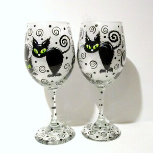 May include: Two clear glass wine glasses with black and white painted designs. Each glass features a black cat with green eyes and a swirling tail. The background of the glasses is white with black dots and swirls.