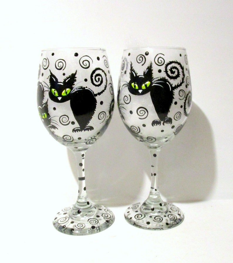 May include: Two clear glass wine glasses with black and white painted designs. Each glass features a black cat with green eyes and a swirling tail. The background of the glasses is white with black dots and swirls.