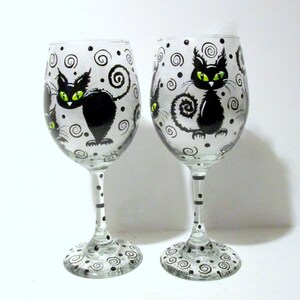 May include: Two clear glass wine glasses with black and white swirling patterns and black cats with green eyes painted on them.