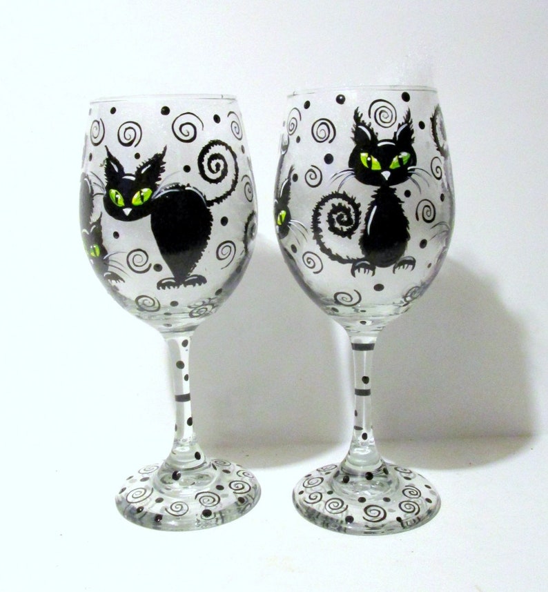 May include: Two clear glass wine glasses with black and white swirling patterns and black cats with green eyes painted on them.