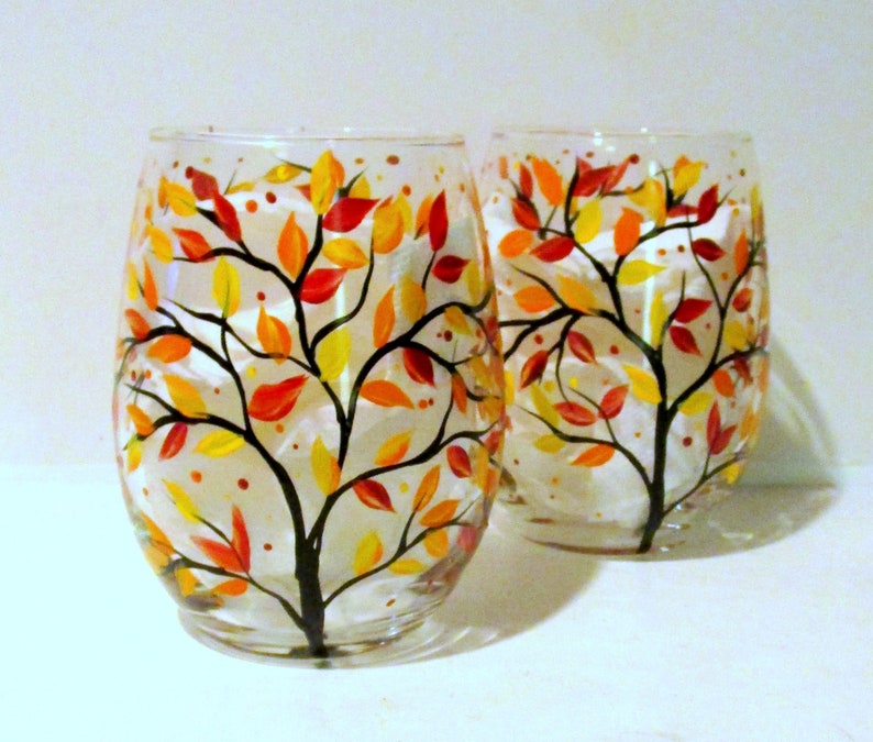 May include: Two clear glass wine glasses with hand painted fall tree designs. The trees have black branches and leaves in shades of red, orange, and yellow. The glasses are on a white surface.