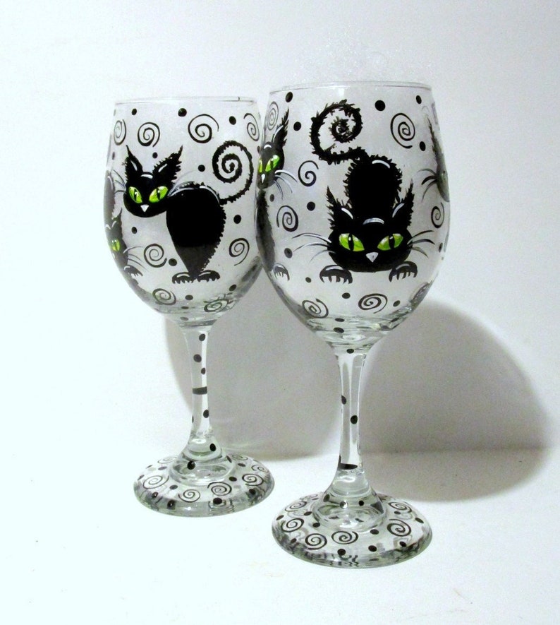May include: Two clear glass wine glasses with black and white painted designs. Each glass features a black cat with green eyes and a swirling pattern.