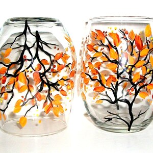 Op de afbeelding: Twee heldere glazen wijnglazen met handgeschilderde herfstbladeren in tinten oranje en geel. De bladeren zijn op het glas geschilderd met zwarte takken.