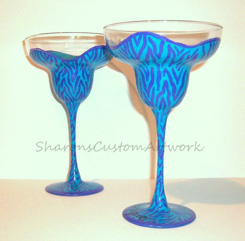 May include: Two clear glass margarita glasses with blue and purple zebra print designs. The glasses are hand-painted and ready to be used for a party.