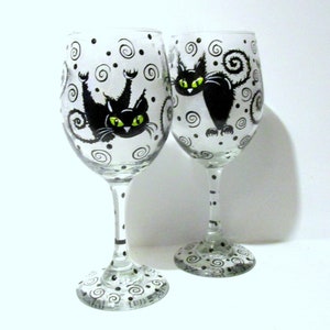 May include: Two clear glass wine glasses with black and white painted designs. Each glass features a black cat with green eyes and a curled tail. The cat on the left glass is upside down.