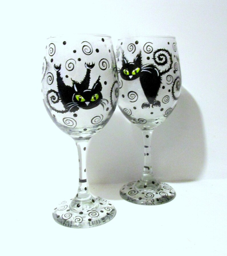 May include: Two clear glass wine glasses with black and white painted designs. Each glass features a black cat with green eyes and a curled tail. The cat on the left glass is upside down.