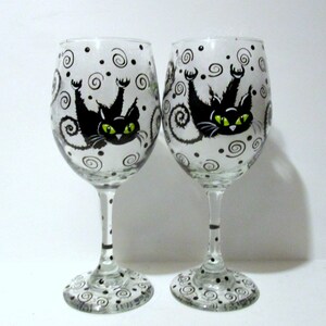 May include: Two clear glass wine glasses with black and white swirling designs and a black cat with green eyes painted on each glass.