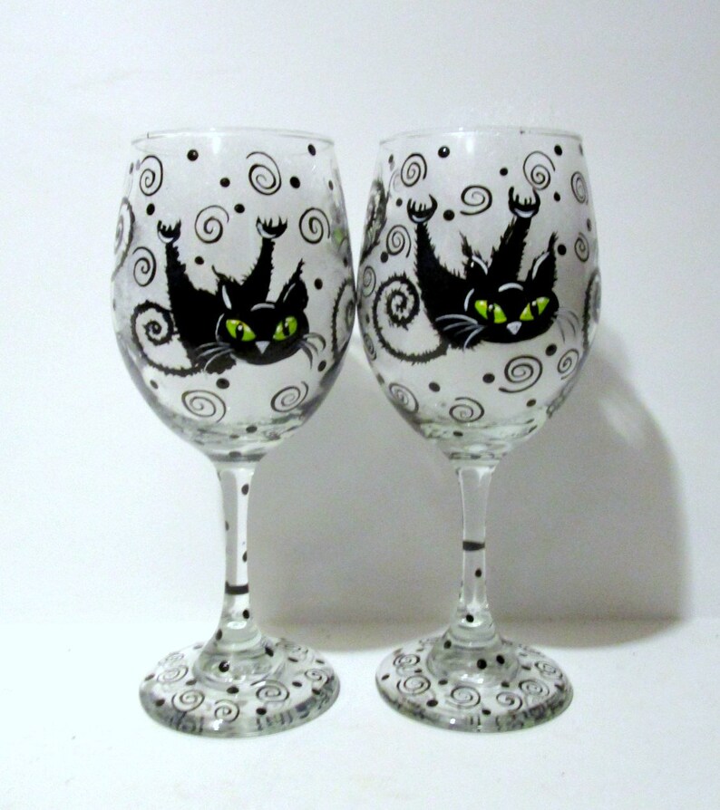 May include: Two clear glass wine glasses with black and white swirling designs and a black cat with green eyes painted on each glass.