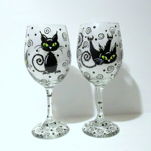 May include: Two clear glass wine glasses with black cats painted on them. The cats have green eyes and are surrounded by black swirls and dots.