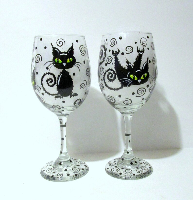 May include: Two clear glass wine glasses with black cats painted on them. The cats have green eyes and are surrounded by black swirls and dots.