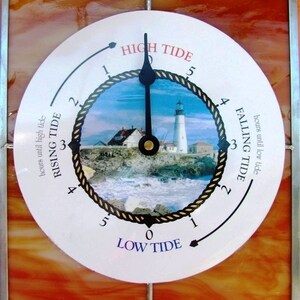 May include: A white circular tide clock with a black hand pointing to high tide. The clock face has a picture of a lighthouse and a rope design around the edge. The text "HIGH TIDE", "LOW TIDE", "RISING TIDE", and "FALLING TIDE" are printed on the clock face. The date and time "2010/11/11 14:11" are printed below the clock.
