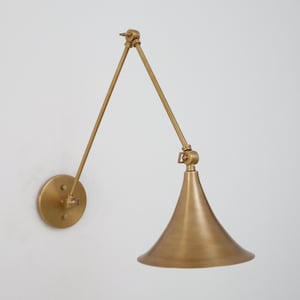 May include: A brass wall lamp with an adjustable arm and a conical shade. The lamp has a circular base and a pivoting arm, enabling directional lighting. The finish has a warm, metallic sheen. The lamp's design is suitable for interior lighting.